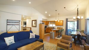 A spacious living area with a blue sofa, wooden coffee table, and a dining table with chairs. A kitchen with wooden cabinets and a chandelier is visible in the background.