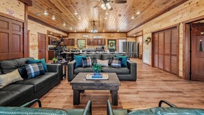 A spacious living area with wooden floors, a ceiling fan, and a kitchen in the background. - Amazing BRAND NEW 8bdrm Cabin-Pool-Arcades-Gazebo (Sevierville)