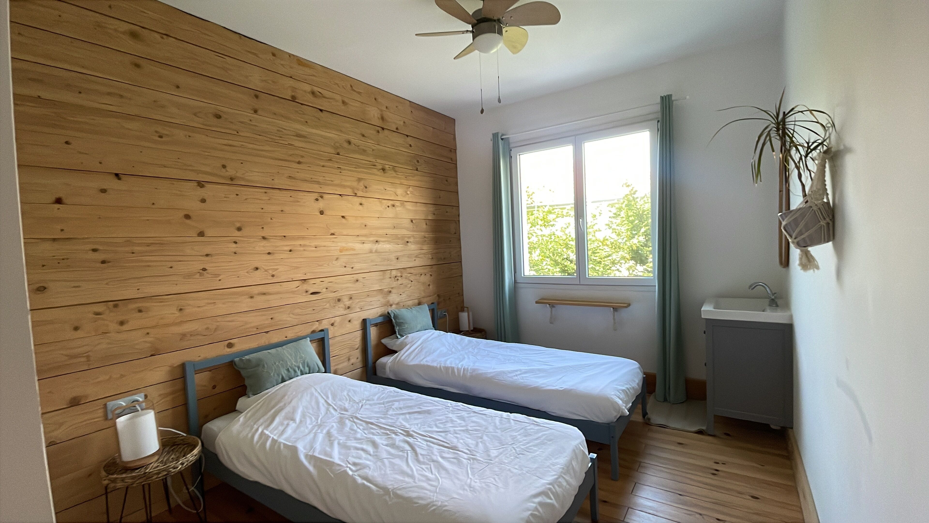 Une chambre avec deux lits, des murs en bois, un ventilateur de plafond, une fenêtre avec des rideaux, une table de chevet avec une lampe et un petit lavabo.