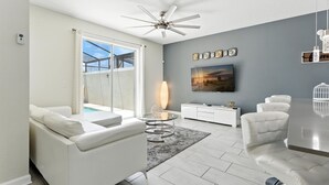 Ein modernes Wohnzimmer mit einem weißen Sofa, einem Glastisch, einem Deckenventilator, einem an der Wand montierten Fernseher und einem durch das Fenster sichtbaren Pool.