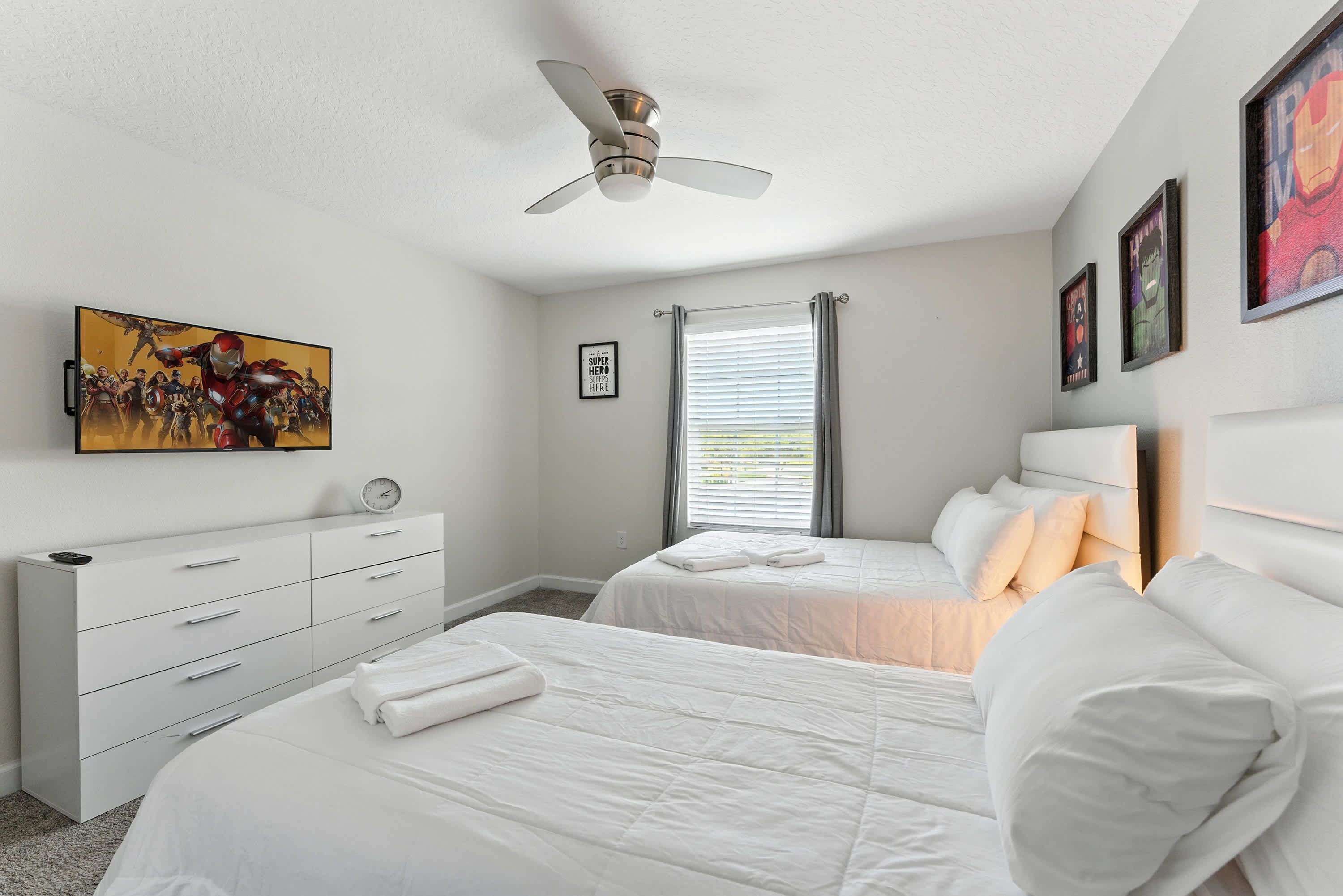 A bedroom with two beds, a ceiling fan, a TV, and artwork on the walls.