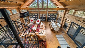 A spacious interior with a high ceiling, wooden beams, a central seating area with a sofa and chairs, a dining table, and a staircase.