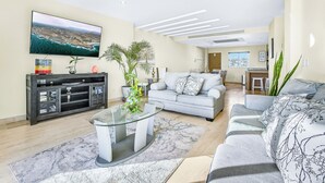 Una sala de estar moderna con una televisión de pantalla plana, una mesa de centro de vidrio, un sofá gris y un área de comedor al fondo.