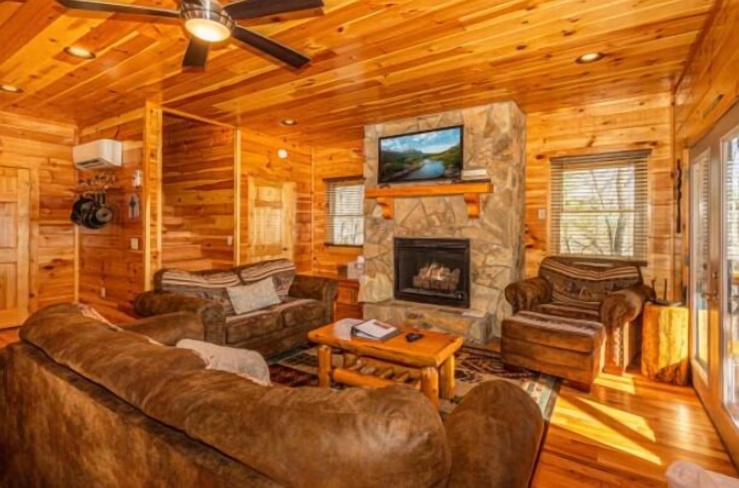 A cozy log cabin interior with a stone fireplace, wooden walls, and a ceiling fan.