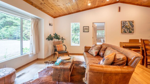 A spacious living room with a wooden ceiling, a leather sofa, a dining table, and a large window offering a view of greenery. - Coastal Redwood Bliss | Little River (Little River)