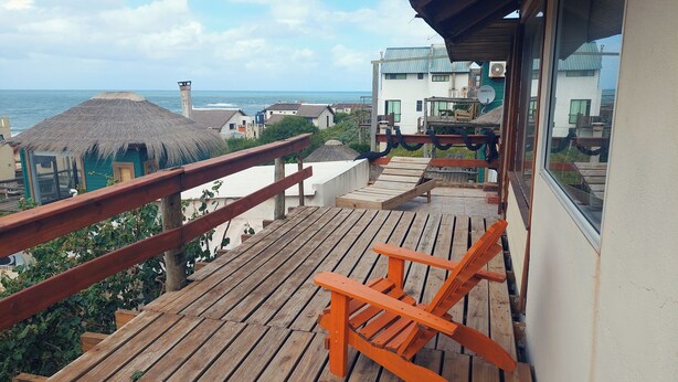 Una terraza de madera con barandilla, una estructura con techo de paja y vista al mar y edificios.