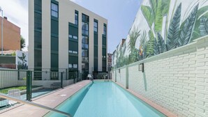 Una piscina con borde de baldosas, rodeada por un edificio con ventanas amplias y un mural de hojas tropicales en la pared.