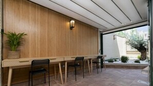 Un patio couvert avec une table en bois et quatre chaises, une plante en pot, et une vue sur un jardin agrémenté d’un mur en briques et de verdure.