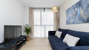 Un salon moderne avec un canapé bleu foncé, un téléviseur à écran plat posé sur un meuble noir, une grande peinture abstraite accrochée au mur et une grande fenêtre ornée de rideaux légers.