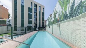 Une piscine avec un bâtiment moderne en arrière-plan, une fresque représentant des palmiers et un mur de briques.