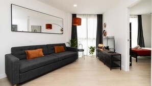 Un salon moderne avec un canapé gris, une table basse et un téléviseur. Un grand miroir orne le mur, et une fenêtre est agrémentée de rideaux.