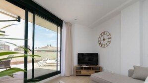 A modern living room with a large sliding glass door leading to an outdoor terrace, a flat-screen TV on a wooden stand, and a minimalist decor.