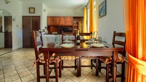 On aperçoit un coin repas avec une table en bois dressée pour quatre personnes, entourée de chaises. Au fond, on distingue une cuisine avec des meubles en bois et un réfrigérateur.