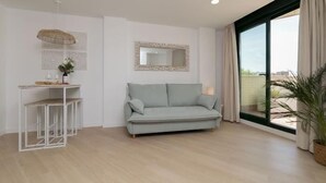 Ein modernes Wohnzimmer mit einem hellgrauen Sofa, einem kleinen weißen Tisch mit Vase und Gläsern, einem dekorativen Spiegel und einem großen Fenster mit Vorhängen.