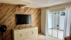 A living room with a flat-screen TV mounted on a wooden wall, a white cabinet, a fan, and a sliding glass door leading to an outdoor area. - HOUSE/21 PEOPLE/POOL/AIR/4 SPACES/BABY/BILLIARDS (Centro Balneário Camboriú)