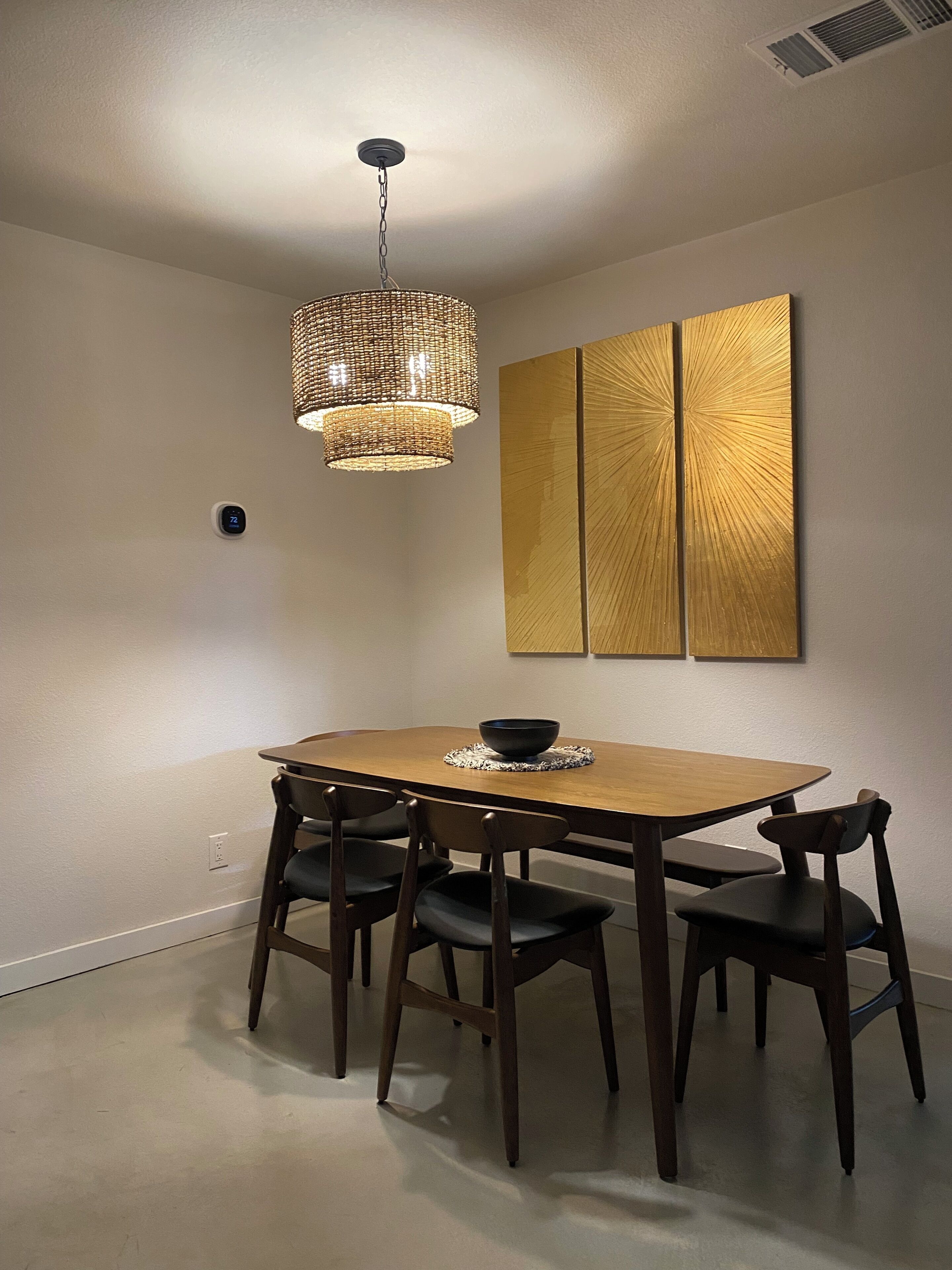 A dining area with a wooden table, four chairs, a woven pendant light, and a wall-mounted decorative panel.