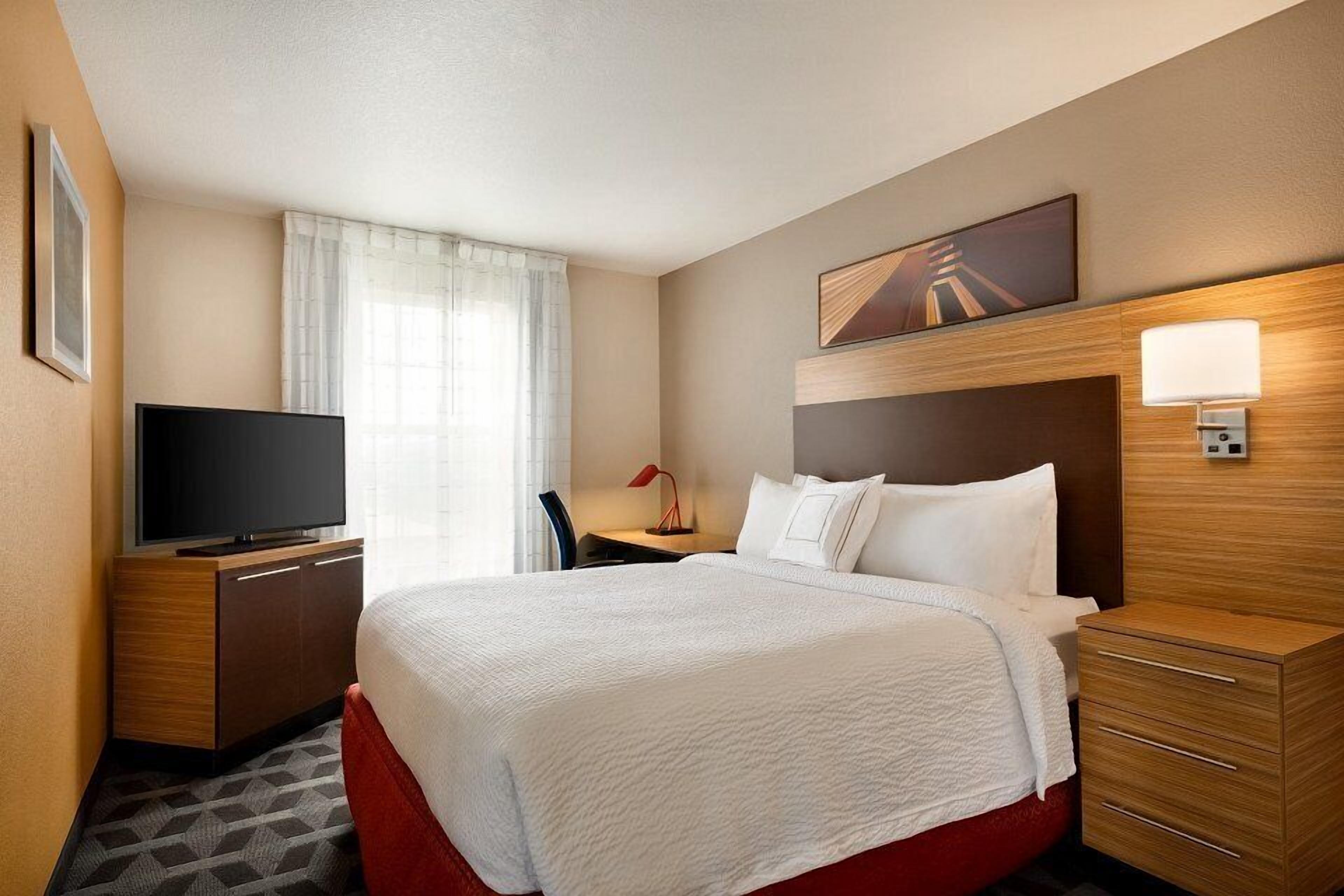 A hotel room with a large bed, a TV on a wooden cabinet, a desk with a chair, and a window with curtains.