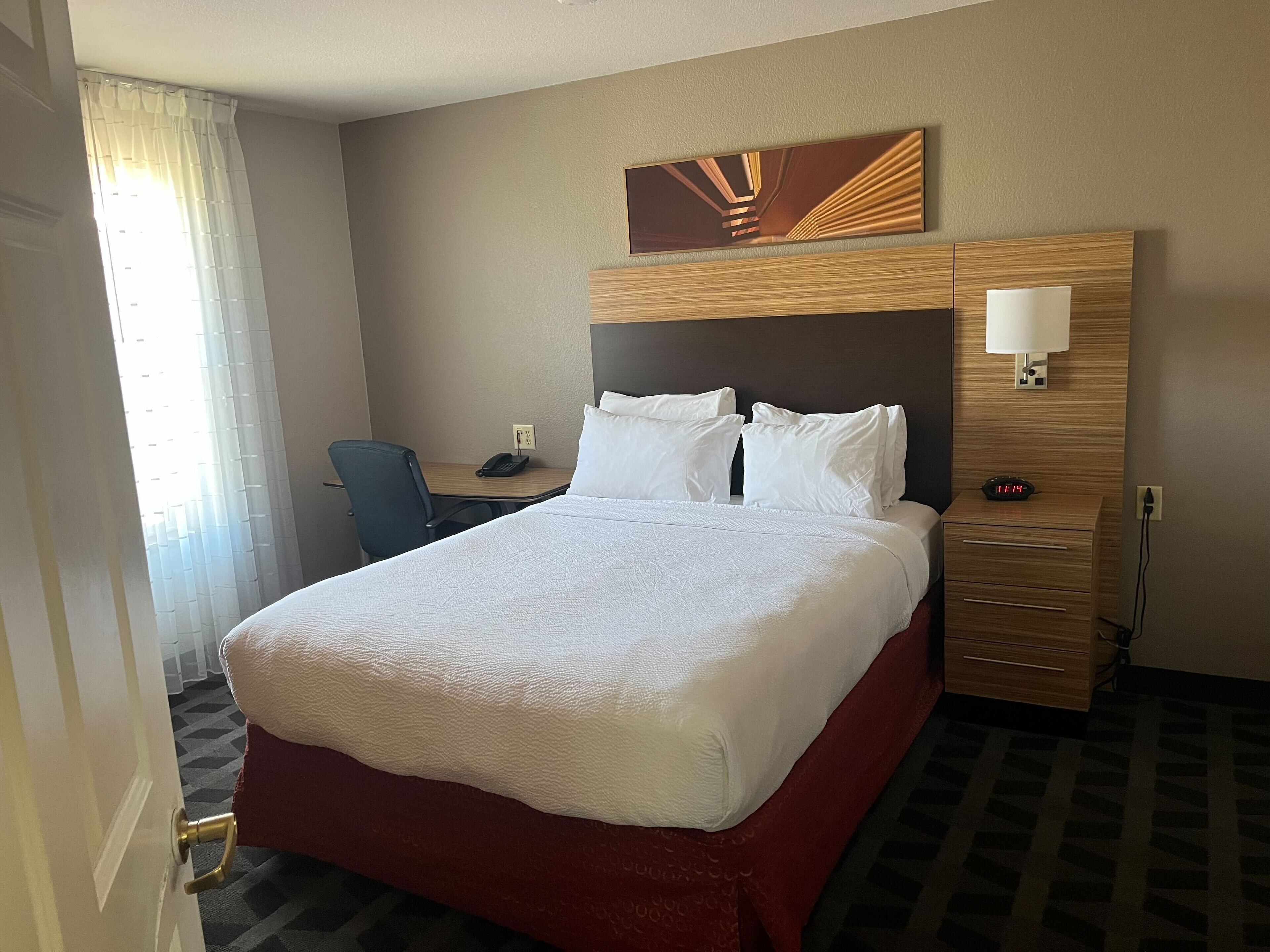A hotel room with a large bed, a desk, a chair, a nightstand, and a wall-mounted lamp.
