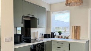 Una cocina moderna con armarios verde claro, horno negro y encimera blanca.  Hay una lámpara colgante de mimbre y una ventana con vistas al exterior.