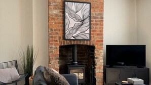 Una sala de estar con chimenea de ladrillo, una obra de arte con marco negro, un sofá gris, un mueble negro para televisor y una mesita.