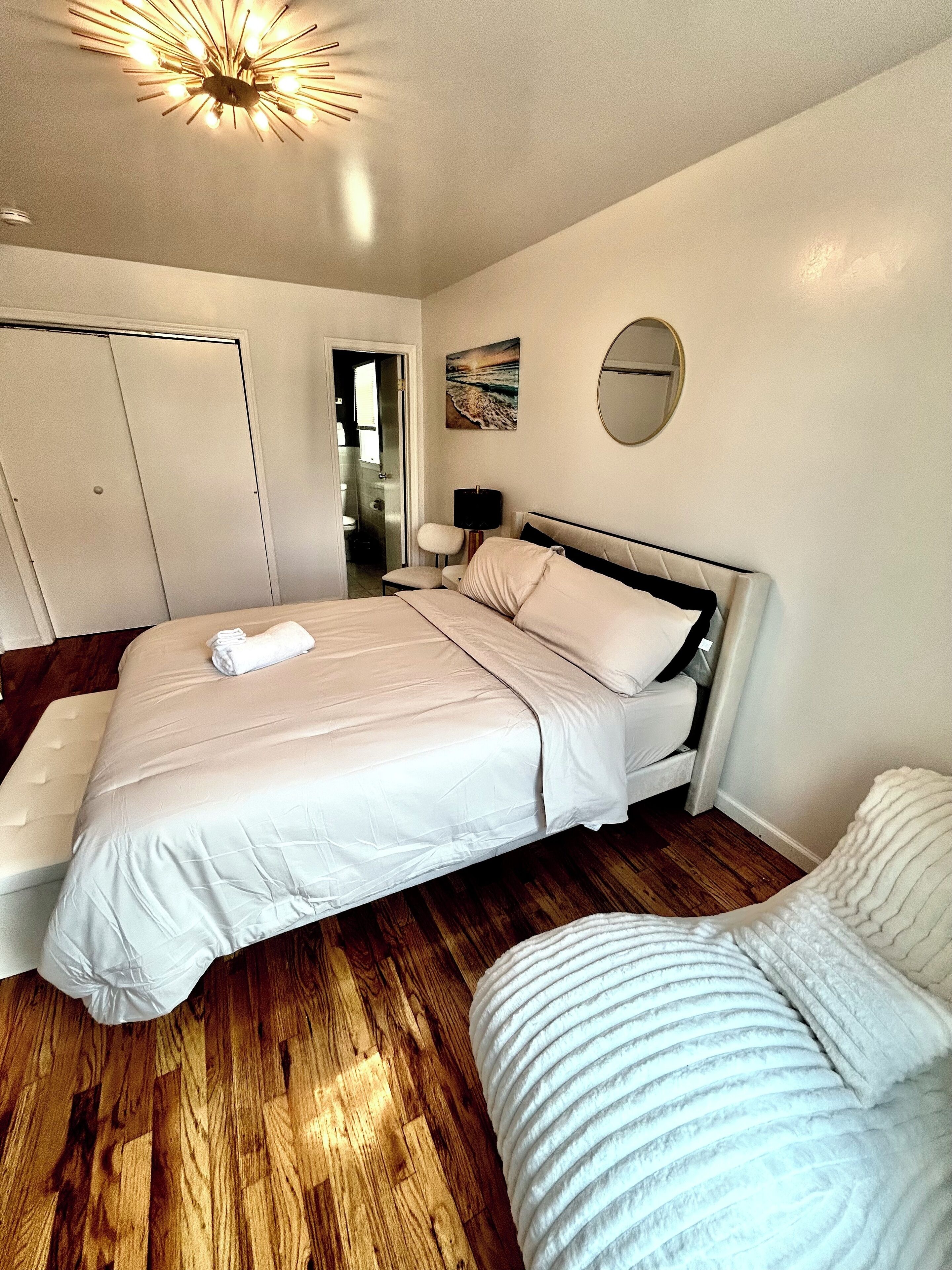 A bedroom with a bed, a chair, a bedside table, a mirror, and a ceiling light fixture.