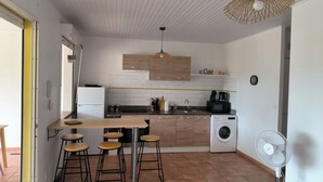 A compact kitchen with a washing machine, a coffee maker, and a fan. There are stools and a wooden table. - Charming T3 apartment in Le Diamant (Le Diamant)