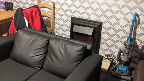 A living room with a black leather sofa, a wooden chair, a small table with a red jacket, and a vacuum cleaner. - Birch Cabin Retreat in Girvan (Girvan)