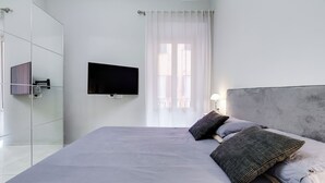 A modern bedroom with a large bed, a flat-screen TV, a glass shower enclosure, and a window with sheer curtains.