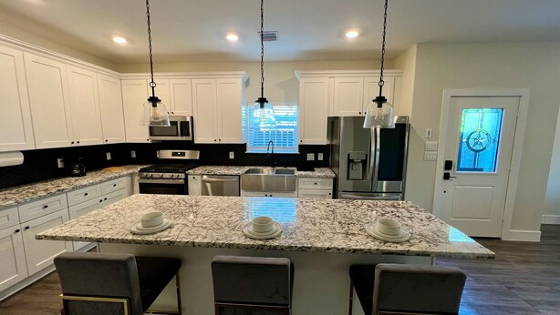 A modern kitchen with white cabinets, granite countertops, a central island with bar stools, stainless steel appliances, and pendant lighting. - Modern Houston Home 7 Min to NRG Stadium & Med Center (Houston)