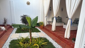 Un jardín cuidado con un elemento vegetal central, rodeado por un camino de grava y césped, que conduce a un espacio de descanso con un sofá y una mesa.