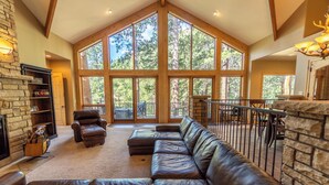 A spacious living area with a large leather sectional sofa, a stone fireplace, and a view of the forest through large windows. - River Bend on Fall River - Riverfront property! (Estes Park)