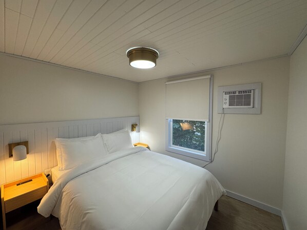 Un dormitorio con una cama, una mesita de noche, una lámpara de pared, una ventana con vistas y un equipo de aire acondicionado.