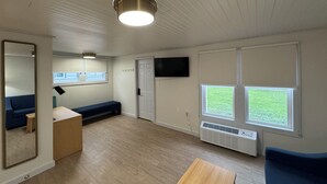 Ein Zimmer mit Holzboden, einem blauen Sofa, einem kleinen Tisch, einem Spiegel, einer Deckenleuchte, einer Tür und einem Fenster mit Blick ins Freie.