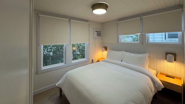 Ein Schlafzimmer mit einem großen Bett, zwei Nachttischen, einer Deckenleuchte und Fenstern mit Jalousien.
