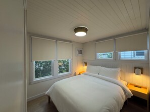 Un dormitorio con una cama grande, dos mesitas de noche, una lámpara de techo y ventanas con persianas.