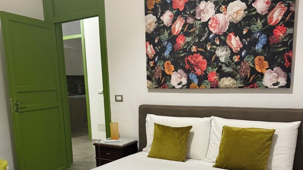 Ein ordentlich bezogenes Bett mit weißen Leinentüchern und zwei Kissen, eine grüne Tür und ein Blumenbild an der Wand.