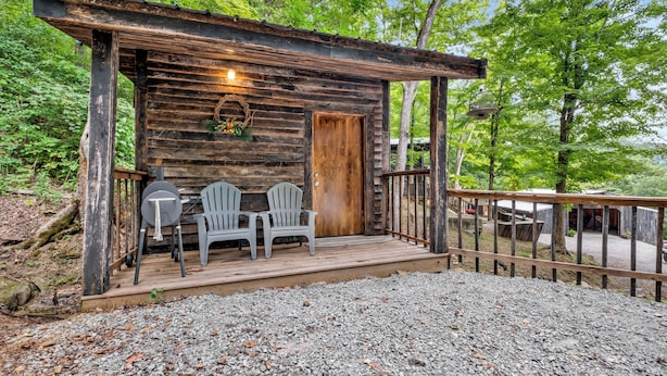A rustic cabin with a wooden porch, two Adirondack chairs, and a gravel ground.