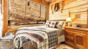 A cozy wooden cabin bedroom with a large window, a bed with a plaid blanket, a wooden dresser, and a hanging lamp.