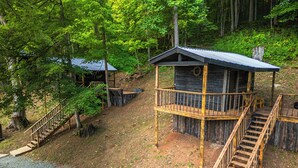A wooden cabin with a balcony and a staircase leading to it, surrounded by trees.