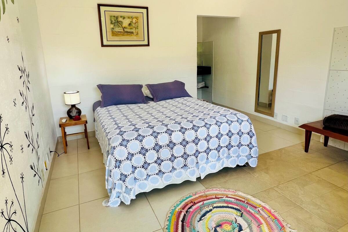 Un dormitorio con una cama, una mesita de noche con lámpara, una alfombra colorida, un cuadro enmarcado en la pared y un espejo.