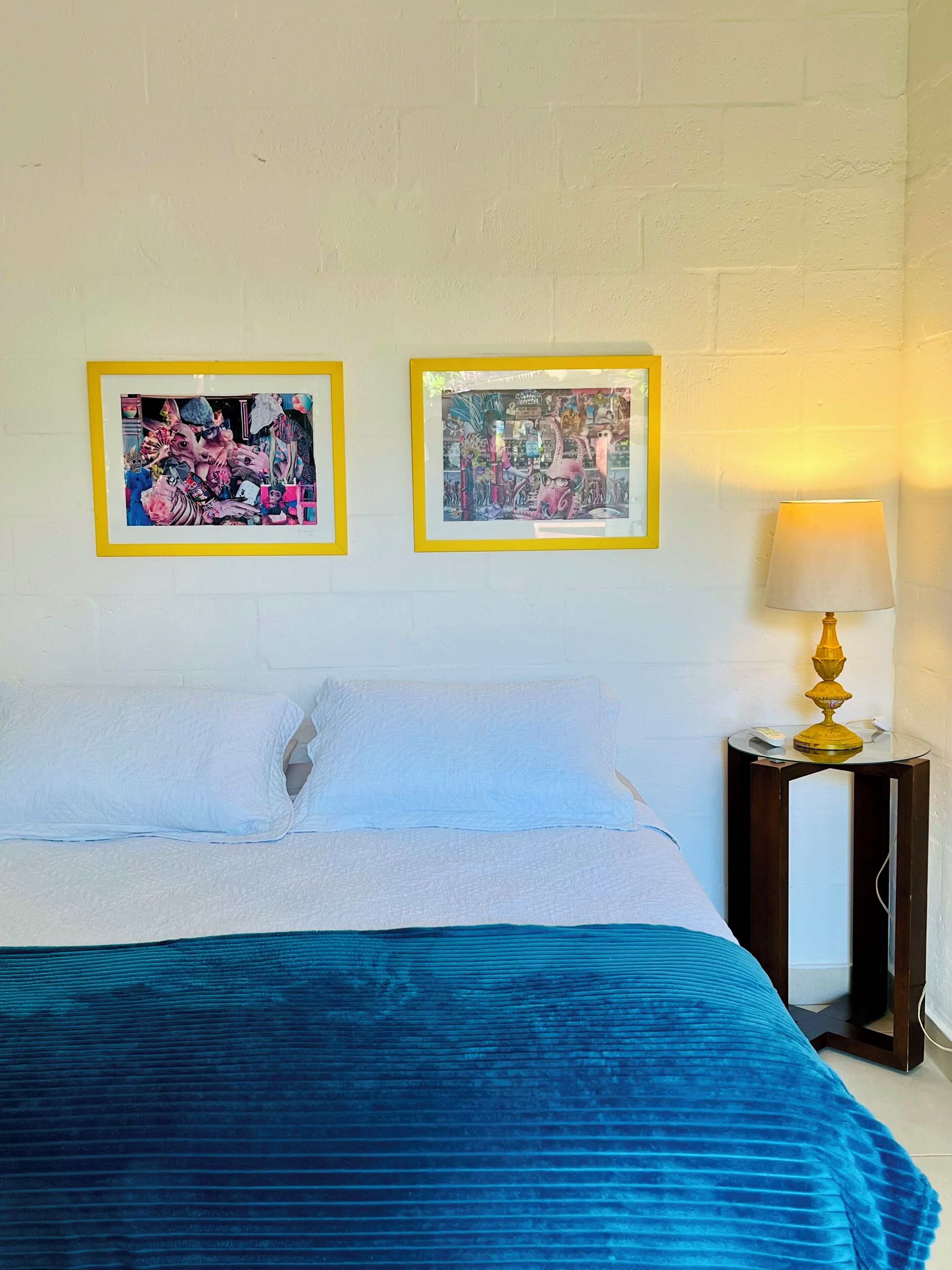 Une chambre d’hôtel avec un lit, deux œuvres d’art encadrées au mur et une table de chevet avec une lampe.