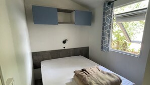 Ein kleines Zimmer mit einem Einzelbett, einer weißen Matratze, einem Kopfteil, einem Fenster mit Blick auf Grün und einem blauen, an der Wand befestigten Regal über dem Bett.