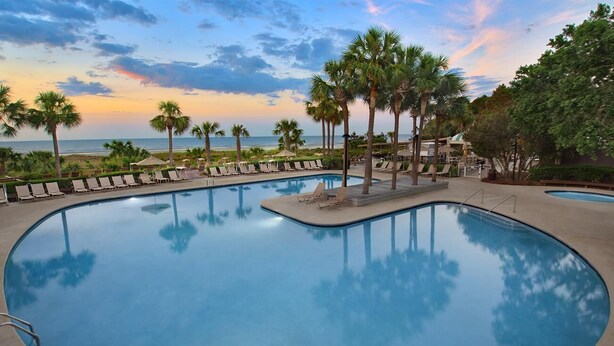 A curved pool with lounge chairs and palm trees, surrounded by a resort area.