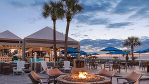 A beachside outdoor seating area with a fire pit, lounge chairs, and umbrellas.