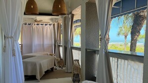 A room with two beds, a large window with a view of the ocean, and a ceiling fan.