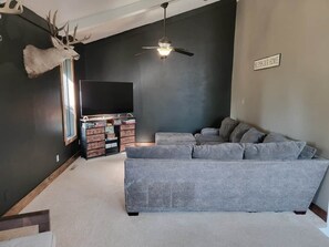 Ein Wohnzimmer mit einem grauen Ecksofa, einem aufgehängten Hirschgeweih, einem Deckenventilator und einem Schild mit der Aufschrift 'Bless Our Home'.