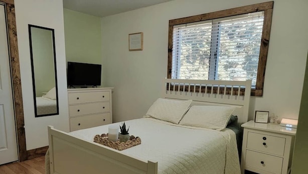 A bedroom with a bed, bedside tables, a mirror, a TV, and a window with blinds.