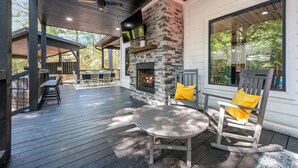 A covered outdoor patio with a stone fireplace, wooden furniture, and large windows.