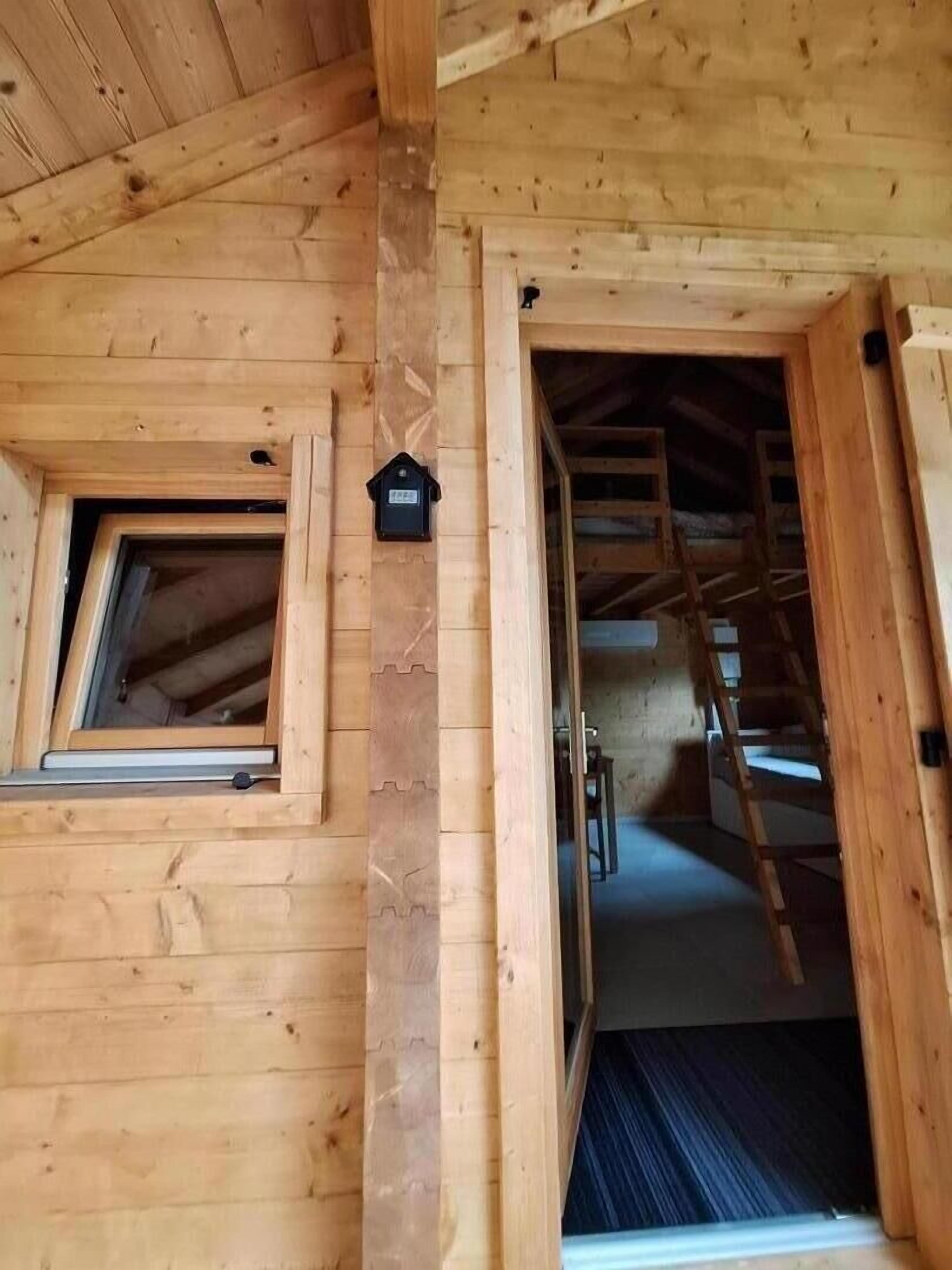 A wooden cabin with a loft bedroom, a window, and a door leading to a staircase.