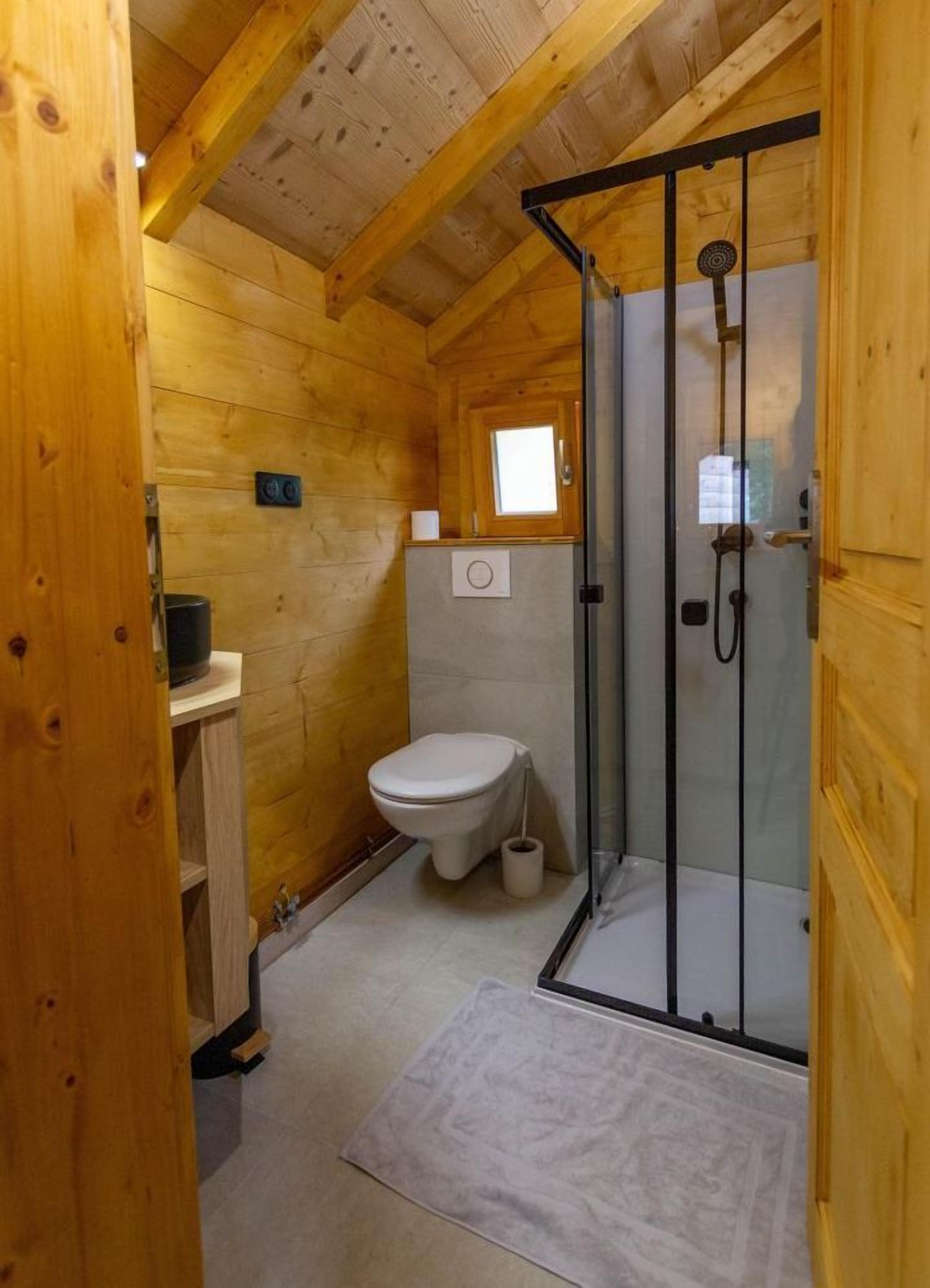 A compact wooden cabin bathroom with a toilet, a shower enclosure, and a small window.